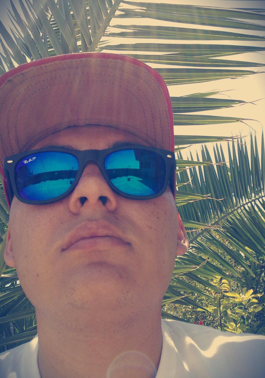 Close-up portrait of Yamil wearing a cap and mirrored sunglasses in front of palm leaves.